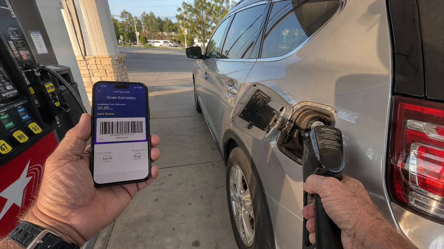 A close-up photo of an older person's hands scanning a fuel loyalty discount barcode from a phone screen at a gas station pump.
