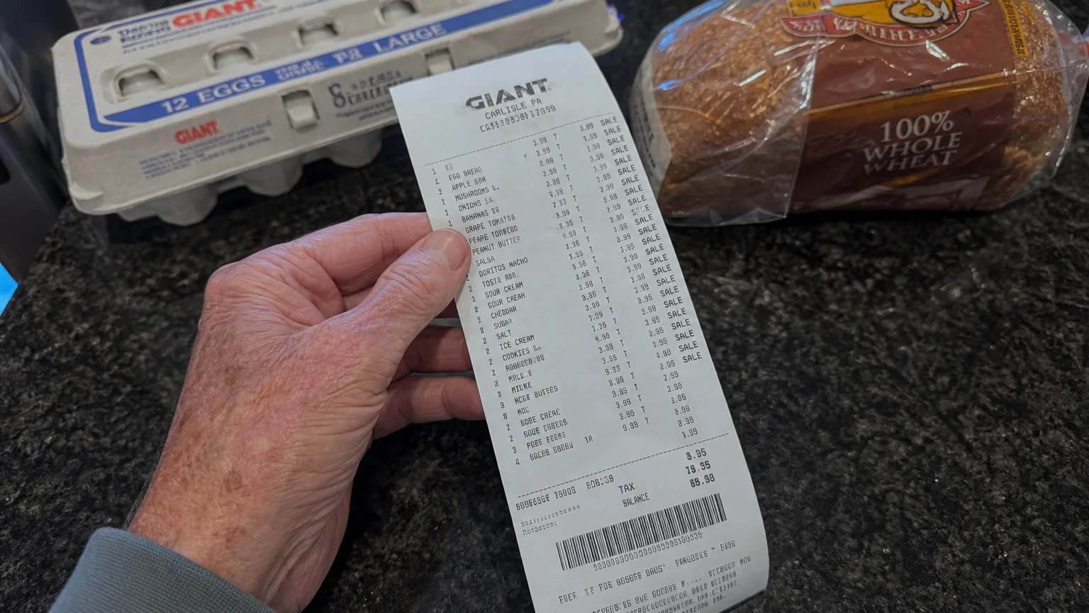 A close-up photograph of an older hand holding a grocery receipt on a kitchen counter next to everyday food items.
